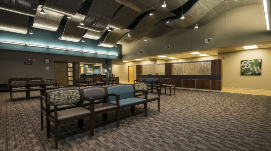 Trinity Health Western Dakota-P-Int2-1024x680-Healthcare Construction Waiting area with shades of brown carpet tiles, brown and teal chairs, check-in desks to the back and wavy ceiling tiles