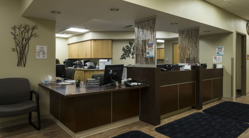 Trinity Health Western Dakota-P-Int3-1024x680-Healthcare Construction Dark brown wood and speckled black countertop medical reception desks with brown and clear glass decorative dividers.