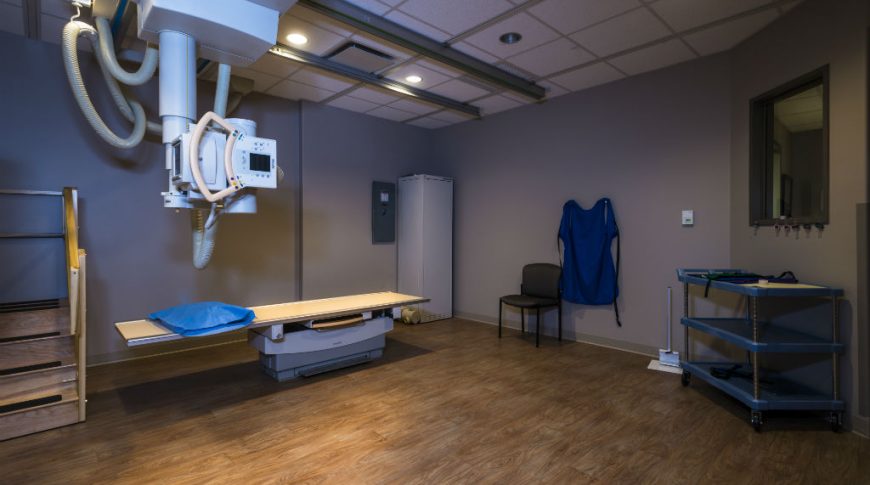 Trinity Health Western Dakota-P-Int4-1024x680-Healthcare Construction X-ray room with faux wood laminate flooring, x-ray machine hanging down from center of ceiling and greige walls