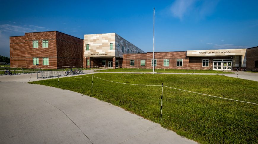 Exterior view of brown brick building with shades of beige panel bump-out entrance sections, one for the elementary school entrance and the other for the middle school entrance. with grassy knoll and sidewalks to the front