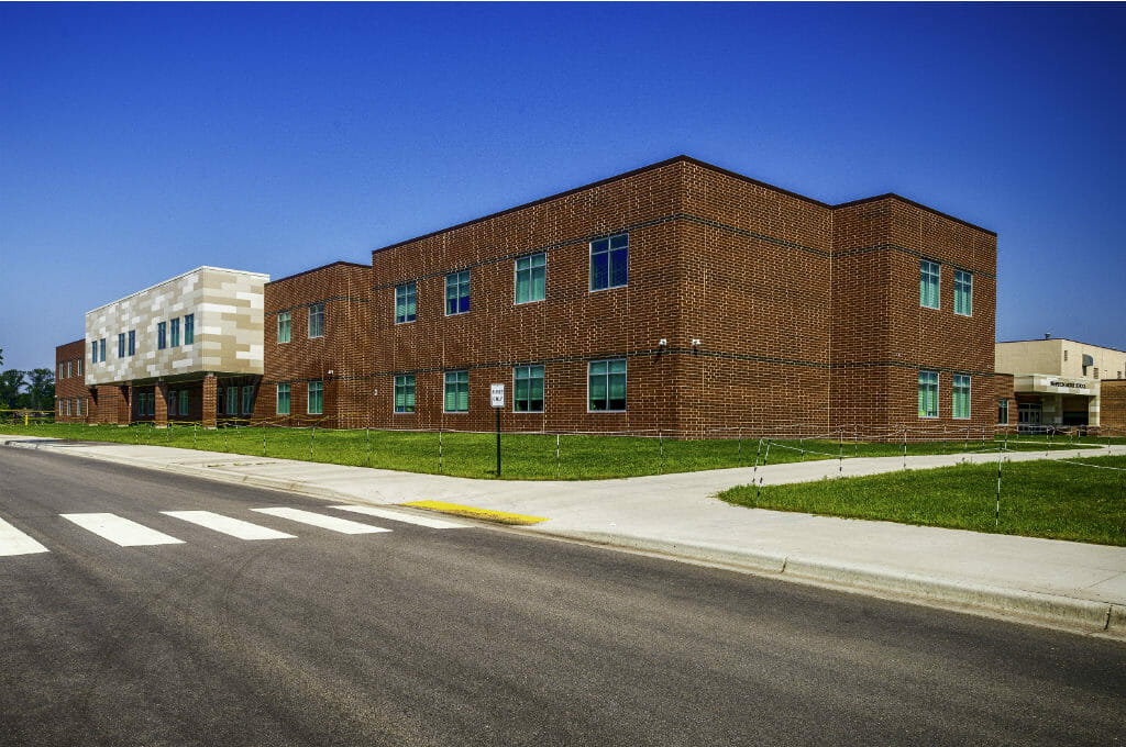 Exterior view from street of dark brown and black accented rectangular brick school building with windows neatly arranged throughout and a lighter shades of beige paneled bump out in the middle