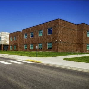 Exterior view from street of dark brown and black accented rectangular brick school building with windows neatly arranged throughout and a lighter shades of beige paneled bump out in the middle