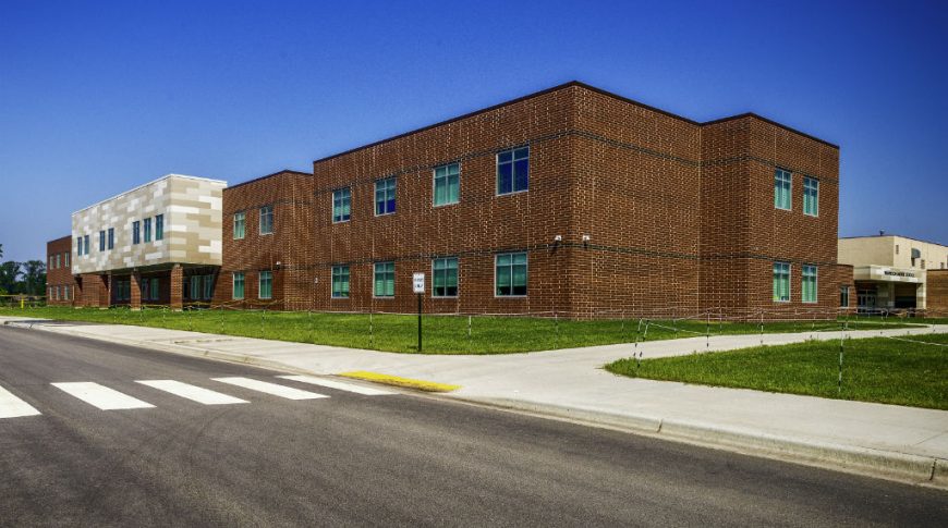 Exterior view from street of dark brown and black accented rectangular brick school building with windows neatly arranged throughout and a lighter shades of beige paneled bump out in the middle