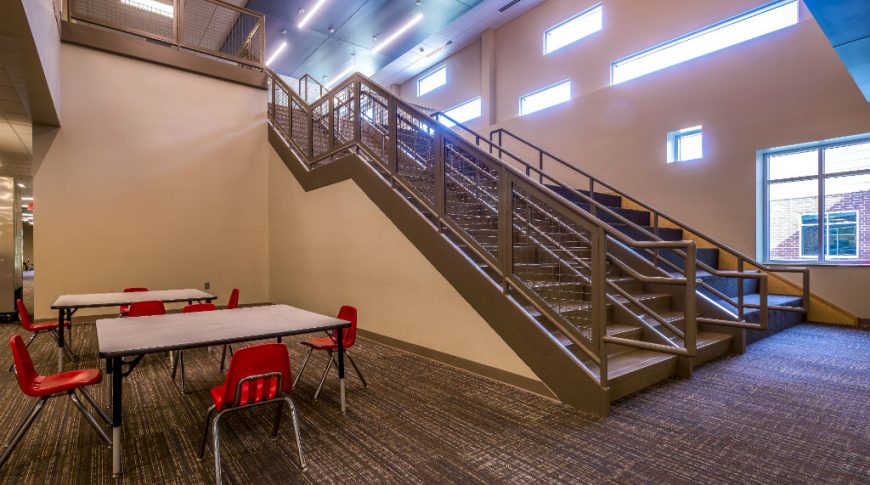 Large double stair case covered in brown vinyl with wall of long rectangular accent windows to the back right side and two small tables with red plastic chairs to the left