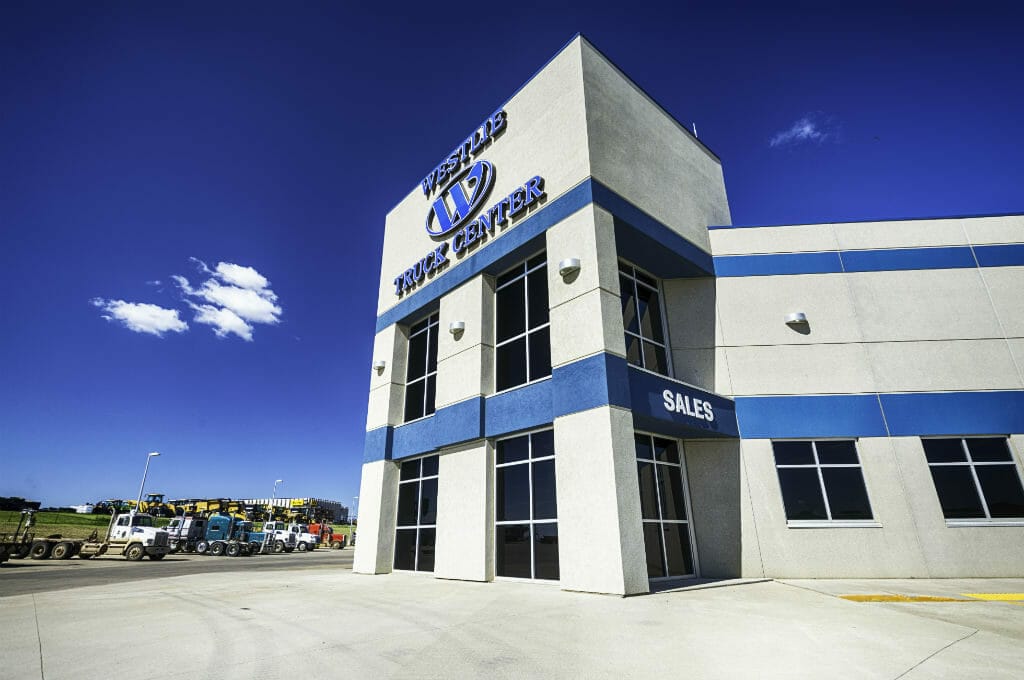 Up-close view of the front outside entrance of the Westlie Truck Center precast beige building with blue stripes in the middle and top with their sign on the top