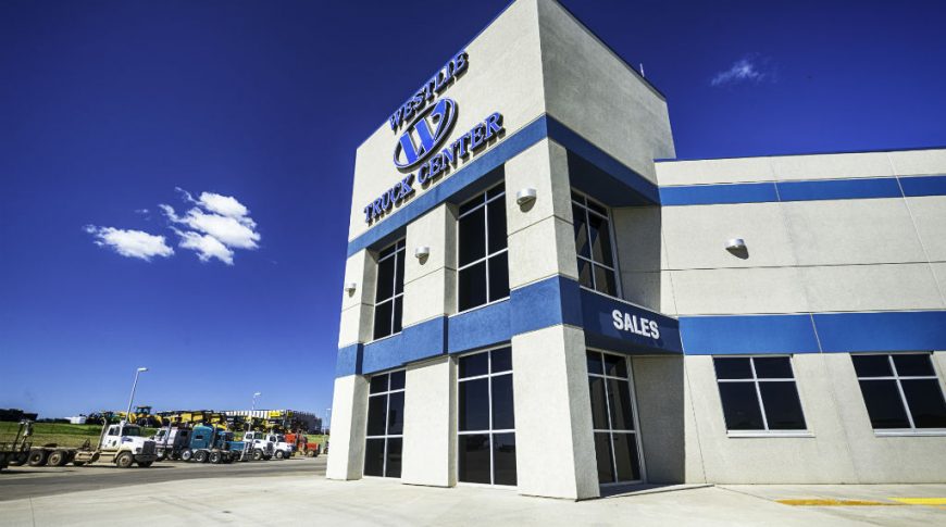 Up-close view of the front outside entrance of the Westlie Truck Center precast beige building with blue stripes in the middle and top with their sign on the top