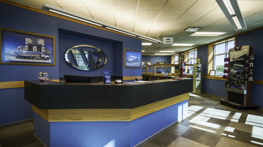 Front desk area with blue colored walls and W mirrored logo for Westlie Truck Center with checkered light and dark beige floor tiles and workstations to the back right