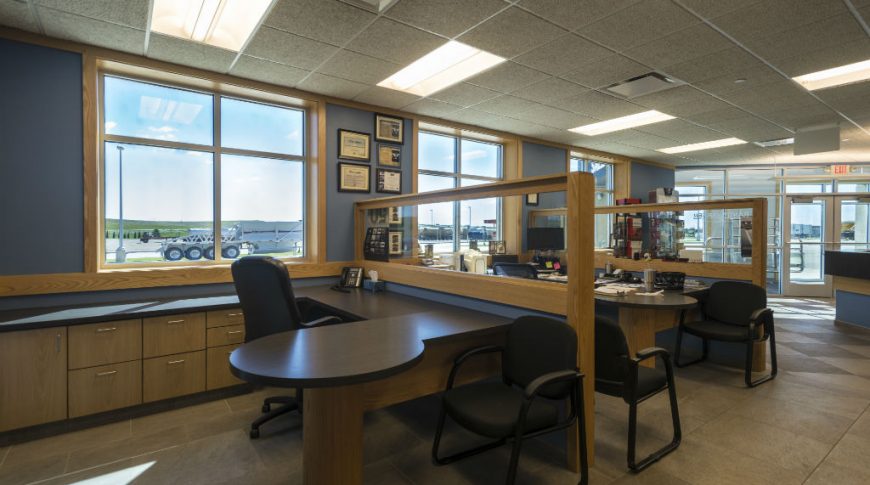 Two desk workstations with light oak cabinetry, glass divider wall and dark countertops with three double windows behind on a blue painted wall.