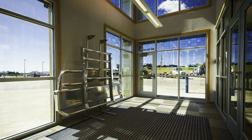 Foyer area with large light and dark beige checkered floor tiles, large built-in grated rug, two-story wiindows and semi-truck grill decorating the back left wall