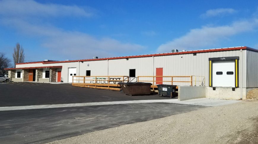 Outside of beige building with red accents with loading bay to the back right and front office entrances to the left.