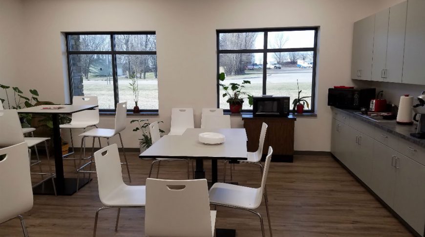 Breakroom with two windows to the back wall, cabinetry to the right wall and tables and chair in the middle.