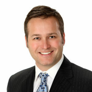 White male with short brown hair and a brown eyes smiling wearing a black suit jacket and white shirt with blue geometric tie on white background.