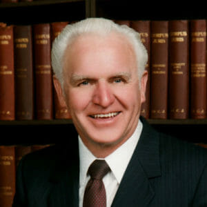 Older white male with white hair smiling wearing a black stripped-suit with white collared-shirt and red polka-dot tie with thesaurus library backdrop.