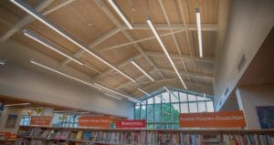 Exposed wood-trussed ceiling in library full of bookshelves, flanked by beige-colored walls and a wall of windows to the rear.