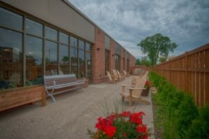 Long, narrow, cedar-fenced outdoor patio with large concrete pebbled pavers, Adirondack chairs and park benches lined with green shrubbery and red geraniums throughout.