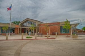 Outside front view of the Fergus Falls Public Library featuring a red brick exterior with a wall of windows at the entrance decorated by planted trees and a flag