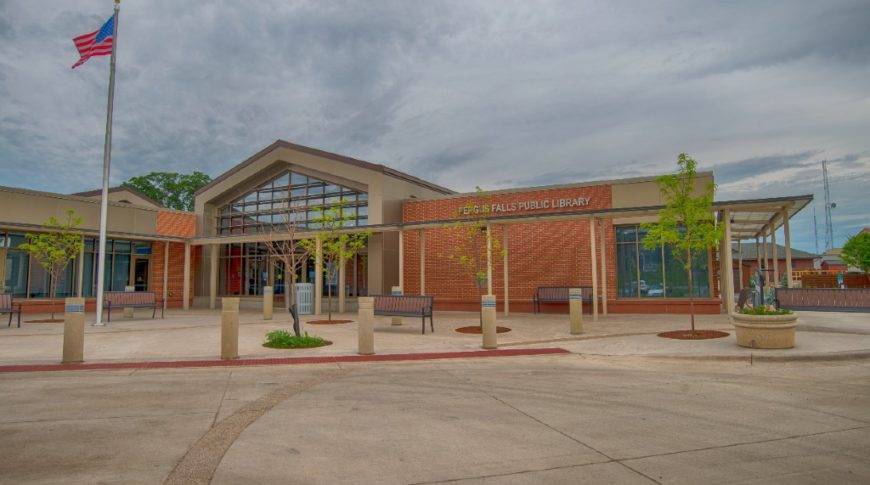 Outside front view of the Fergus Falls Public Library featuring a red brick exterior with a wall of windows at the entrance decorated by planted trees and a flag