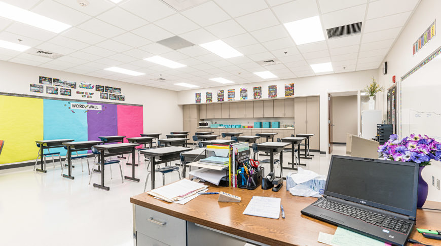 Beige-colored classroom with medium-beige cabinets on the back wall, desks in the middle of the room and a teachers desk in the foreground.