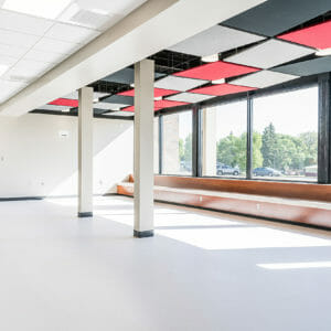 Open commons area with big, bright windows letting sun shine in, a wooden bench across the window sills and school colors in red, black, and grey paneling on the ceiling