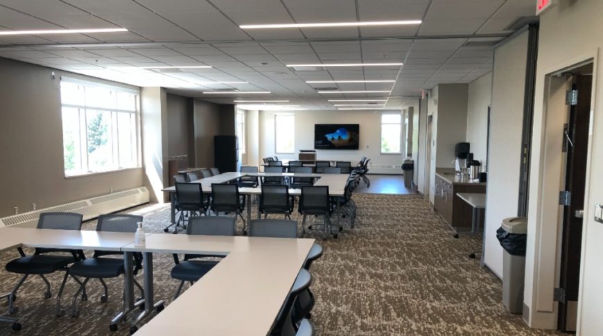 Large rectangular meeting room with tables and chairs grouped in three square areas. There's a mini-bar area to the right of the room with a large tv screen at the far back wall and windows to the far back wall and outside wall. Shades of neutral brown colors blanket the walls, tables, and carpet of the room.