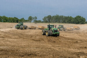 Three green tractors in a dirt field pulling earthwork implements behind