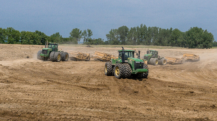Three green tractors in a dirt field pulling earthwork implements behind