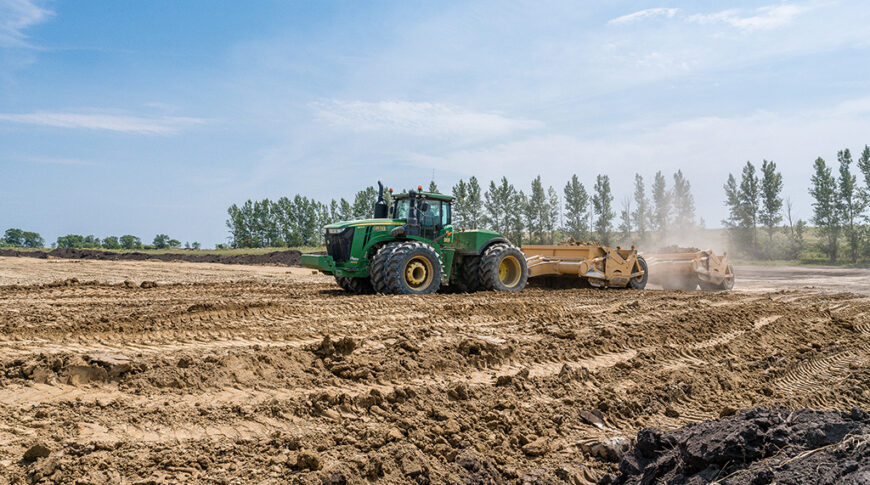 Green tractor pulling dirtwork implement in bare dirt field
