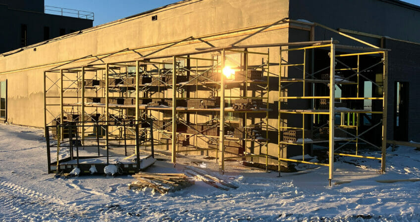 Scaffolding wrapping around the corner of a brick building on a snowy, sunny morning.