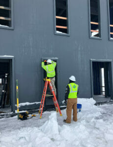 Two men outside shell of building in yellow safety vests and white hard hats. One is atop a ladder and the other on the ground. Snow is beneath both. 