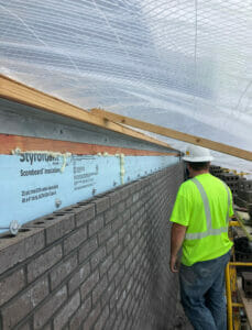 Man in yellow safety shirt standing by partially finished brick outer wall with tarp overhead for colder winter construction months.
