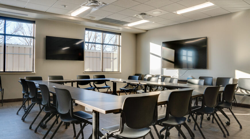 Beige conference room with two windows to the back wall, two large screen tv's to the far back left and right walls, and four large conference rectangular tables put together in a square with opening in the middle and chairs all around.