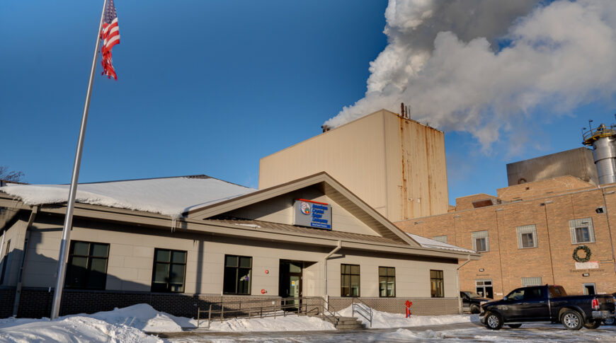 Outside winter view of the new American Crystal Sugar office building. Plant expelling moisture in the background with tall American flag flagpole in the left foreground.