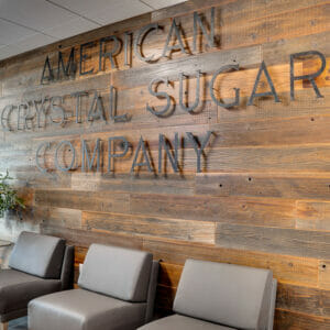 Warm-stained wood paneled wall with a raised metal sign that says "American Crystal Sugar Company" with three gray sleek chairs below and two side tables on either side, one with green plant.