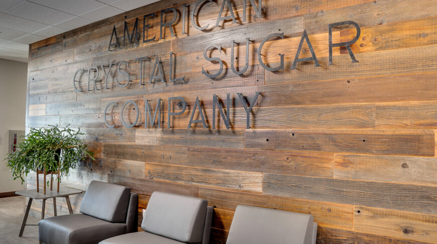 Warm-stained wood paneled wall with a raised metal sign that says "American Crystal Sugar Company" with three gray sleek chairs below and two side tables on either side, one with green plant.