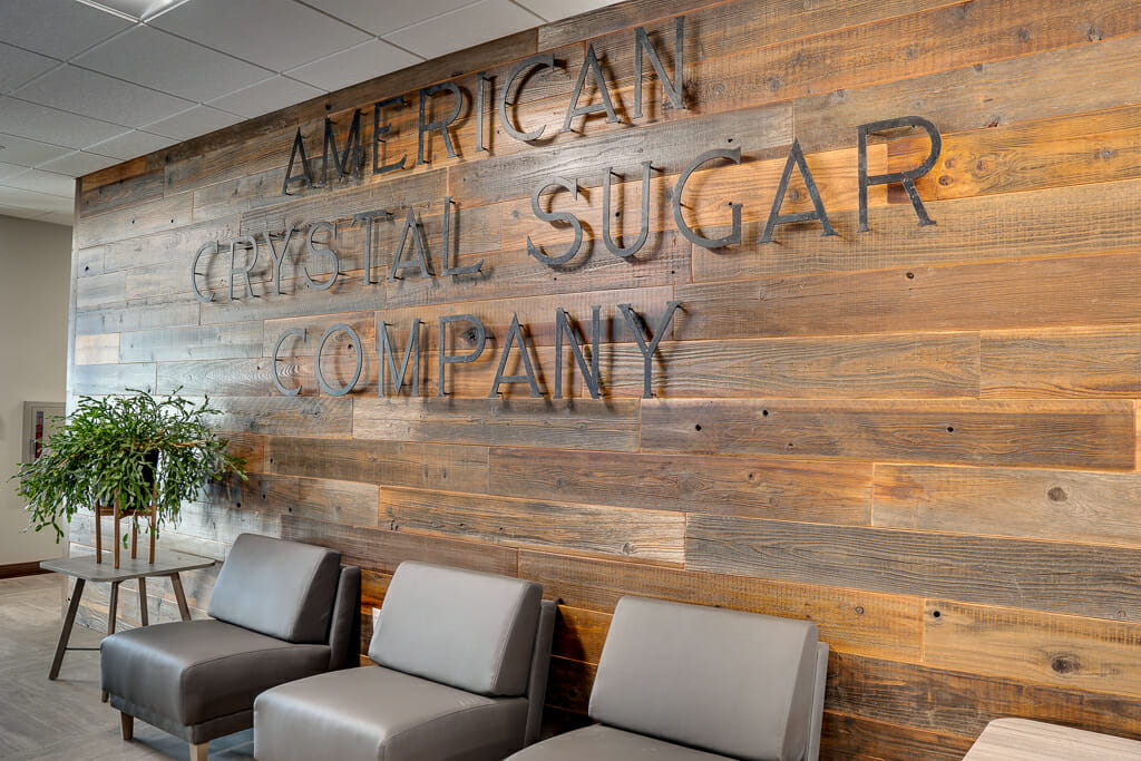 Warm-stained wood paneled wall with a raised metal sign that says "American Crystal Sugar Company" with three gray sleek chairs below and two side tables on either side, one with green plant.