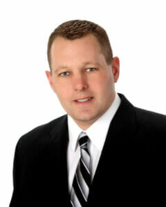 Middle-aged white man with short light brown hair and blue eyes with small smile wearing a black jacket, white button-up shirt and striped black, gray and white tie.