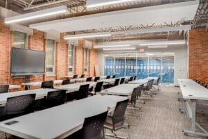 Retrofitted classroom space with red brick exterior and interior walls, exposed metal ceiling with modern lights and long round rectangular tables and chairs spanning the length.