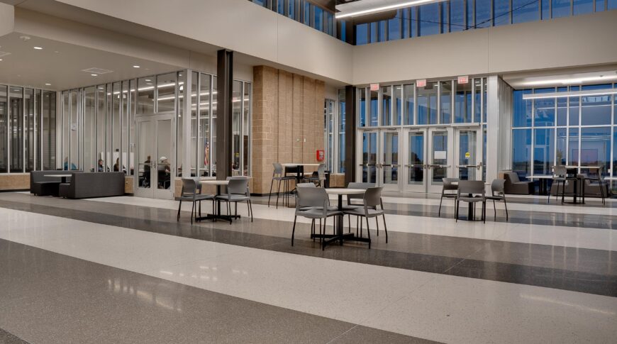 Entryway commons area of high school building with white, gray and dark gray striped floors, and small round tables and chairs throughout.