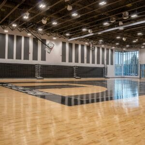 High school gymnasium with bright shiny hardwood floors and black painted basketball court details. Black bleachers folded up on back wall flanked by two-story windows on either side.