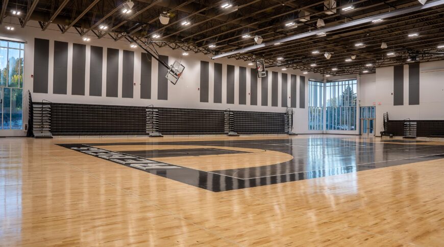 High school gymnasium with bright shiny hardwood floors and black painted basketball court details. Black bleachers folded up on back wall flanked by two-story windows on either side.