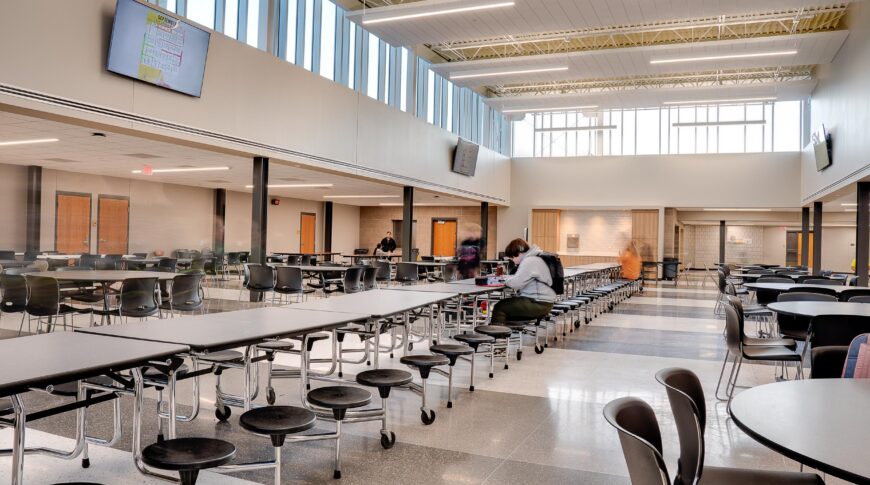 Cafeteria with atrium of windows lining the second story, a row of long rectangular cafeteria tables down the middle with large round tables and chairs on either side.