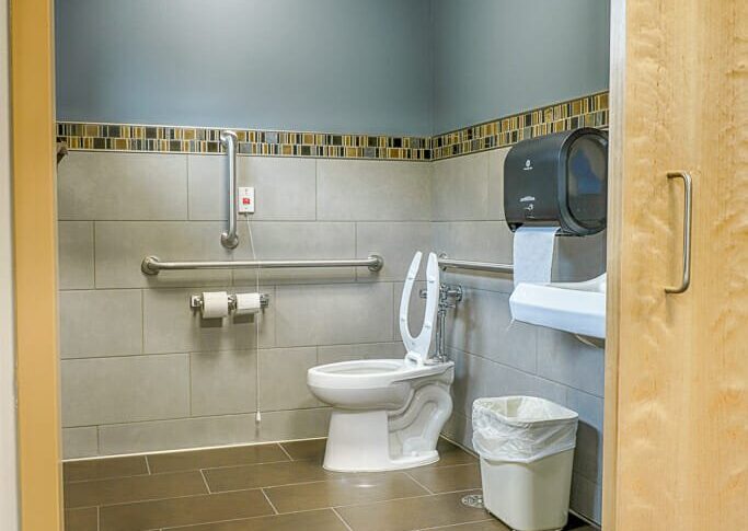 Hospital bathroom with sliding barn maple door leading to brown tiled bathroom with ADA accessible toilet and sink.