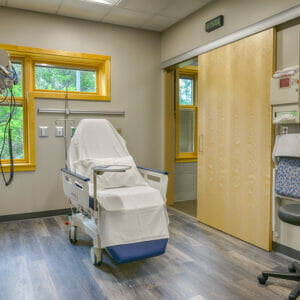 Procedure room with patient chair, sliding maple door to the back right and L-shaped window to the left.