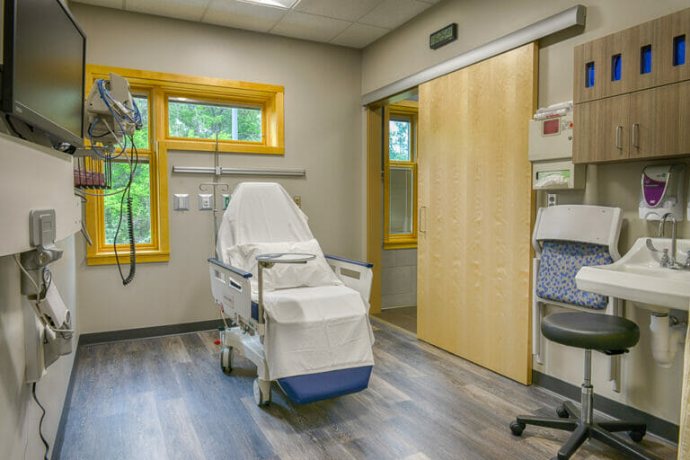 Procedure room with patient chair, sliding maple door to the back right and L-shaped window to the left.