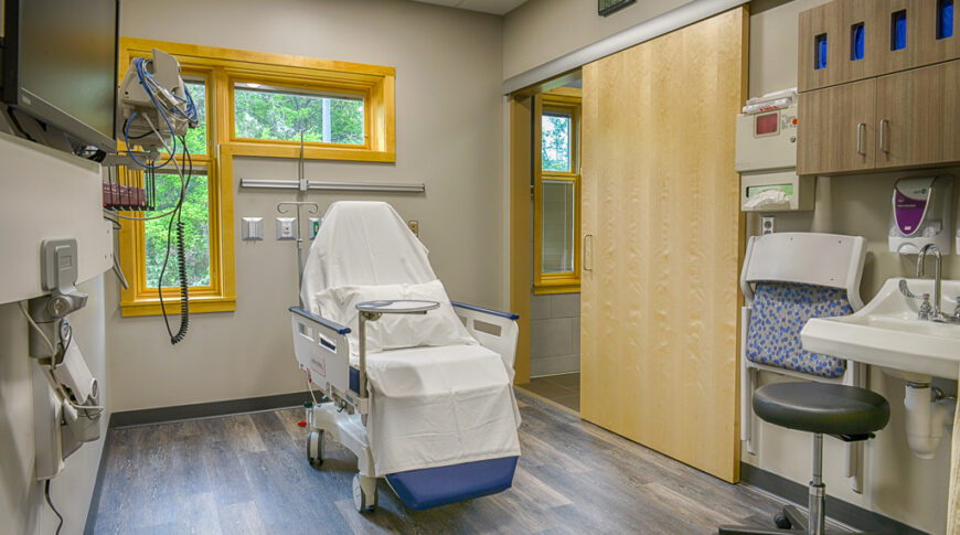 Procedure room with patient chair, sliding maple door to the back right and L-shaped window to the left.