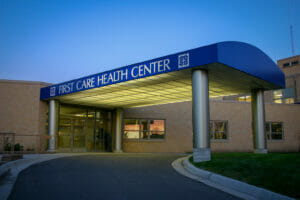 Blue arched covered drive-up with the words "First Care Health Center" on the side and beige bricked building to the back.