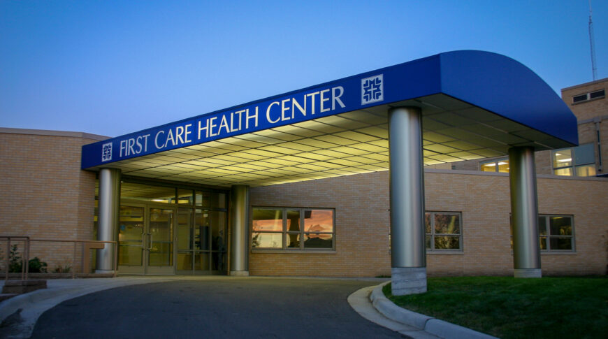 Blue arched covered drive-up with the words "First Care Health Center" on the side and beige bricked building to the back.