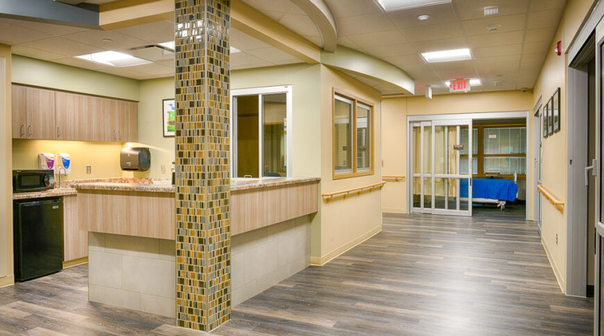 Hallway in hospital area with patient rooms to the right and back walls and a convenience station with mini fridge, microwave and sink to the left.