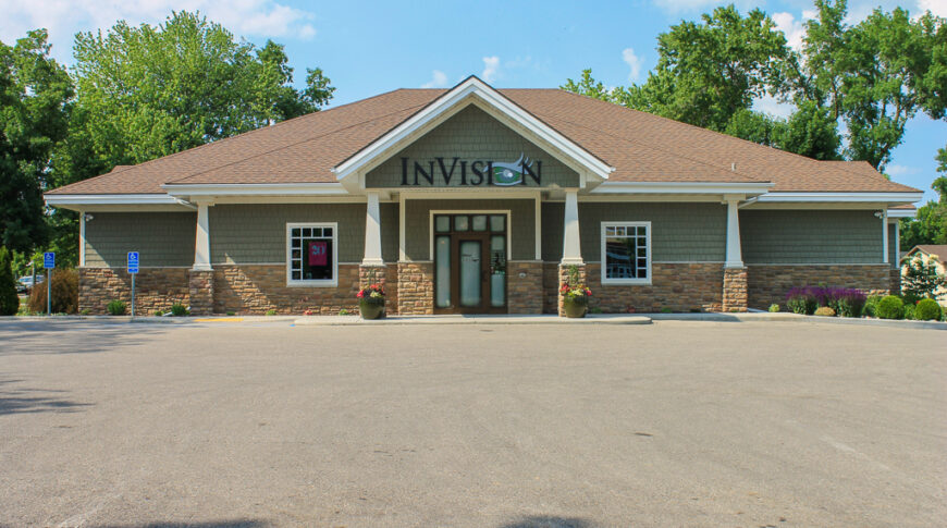 Parking lot exterior view of InVision eye clinic with green shaker siding, white and stone columns and brown shingled roof.