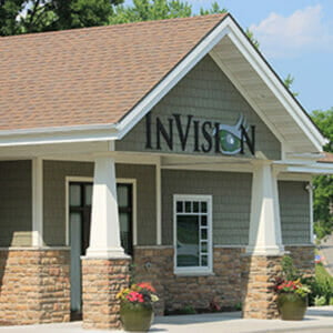 Exterior view of InVision eye clinic with green shaker siding, white and stone columns and brown shingled roof.